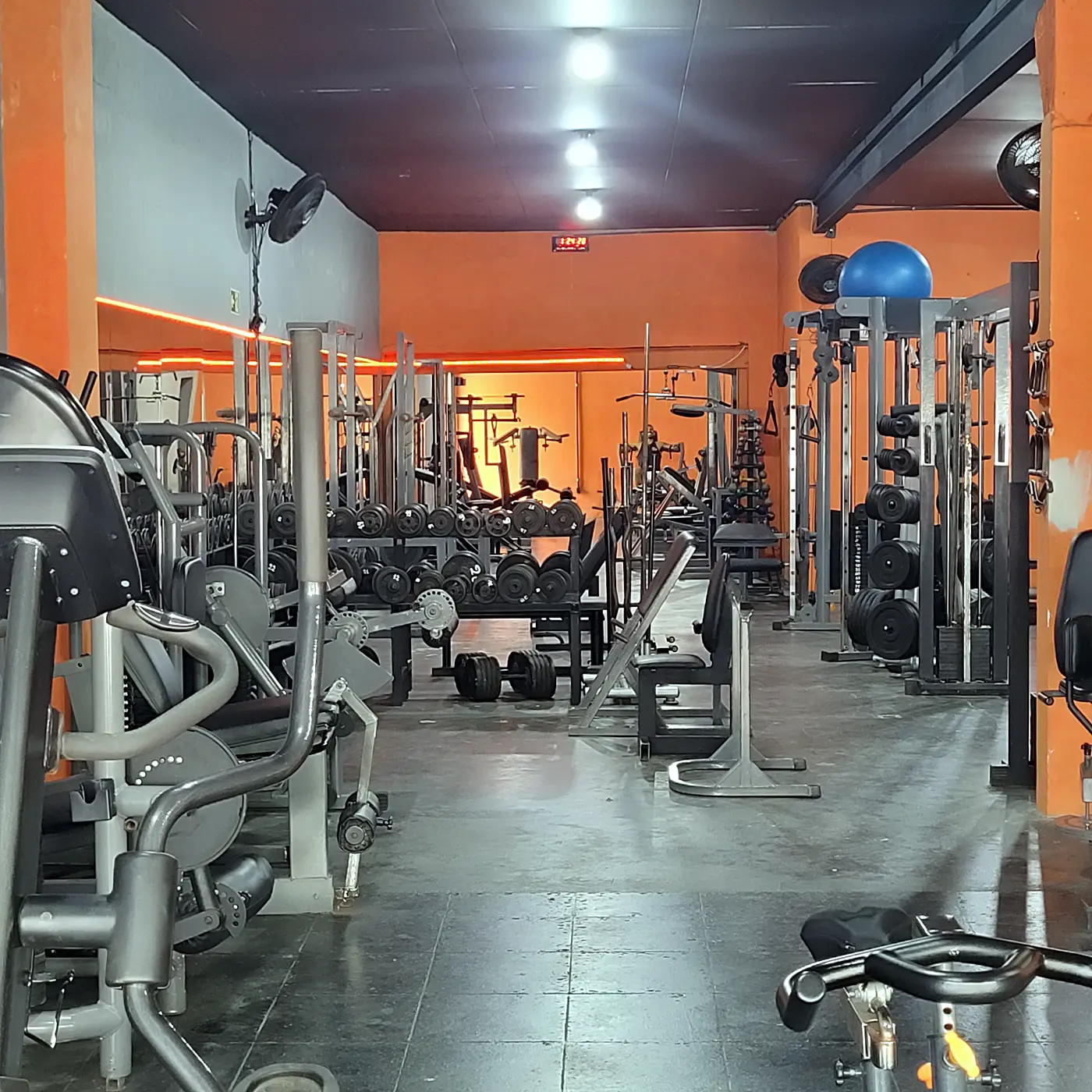 Máquinas e rack de halteres na musculação com parede laranja ao fundo